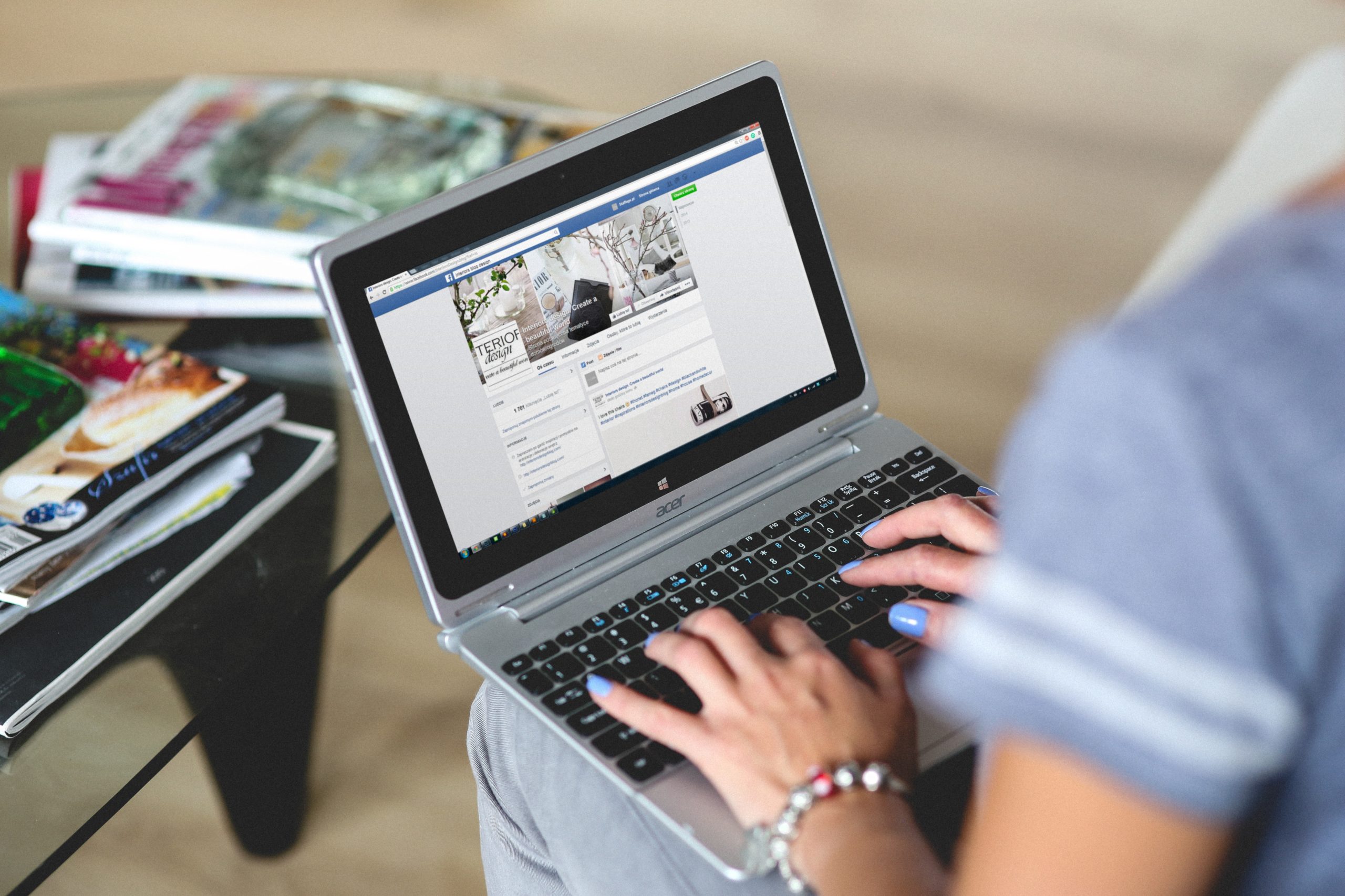 woman with laptop computer on lap looking at Facebook business page with magazines on coffee table in the background; blog: how to conduct a social media competitor analysis